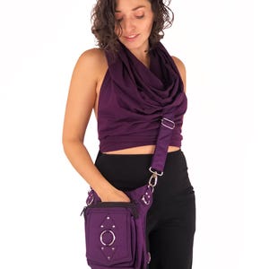 May include: A purple fabric fanny pack with a silver ring and studs. The bag has a zippered pocket and a strap that can be worn over the shoulder or across the body.