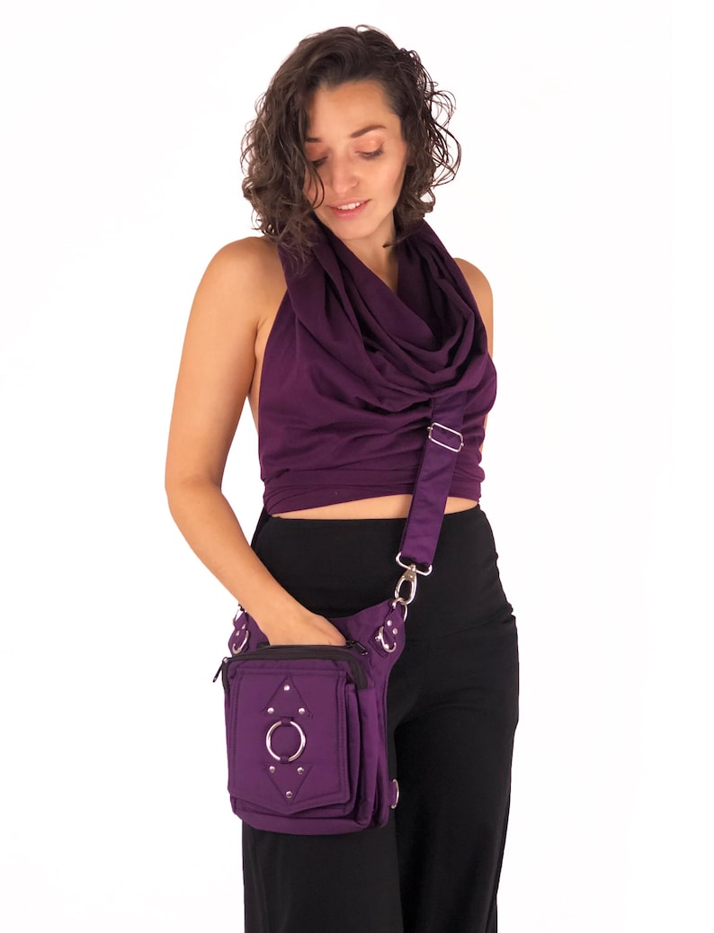 May include: A purple fabric fanny pack with a silver ring and studs. The bag has a zippered pocket and a strap that can be worn over the shoulder or across the body.