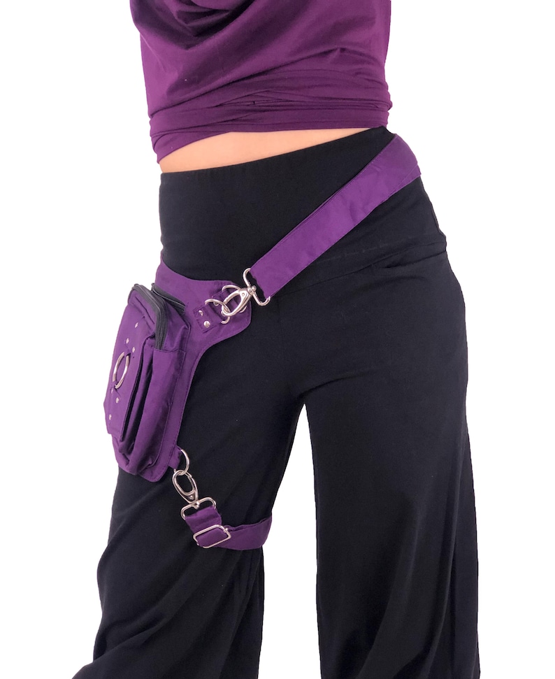 May include: Purple fanny pack with a silver buckle and chain. The pack is worn on the hip and has a strap that goes over the shoulder.