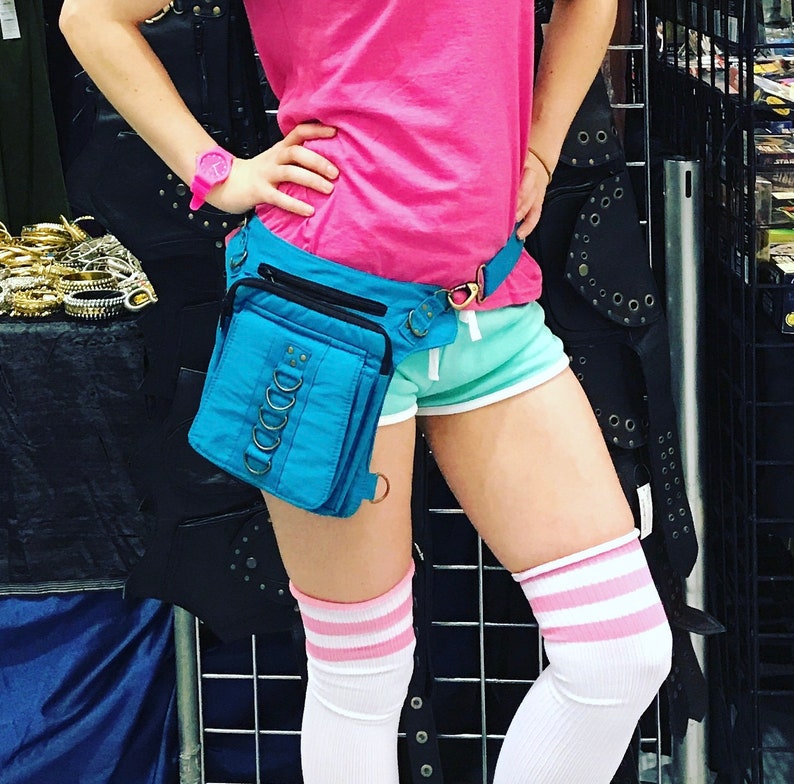 May include: A blue canvas fanny pack with metal rings and a black strap. The fanny pack is worn around the waist of a person wearing a pink shirt, blue shorts, and white socks with pink stripes.