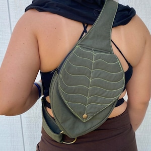 May include: A green canvas crossbody bag with a leaf-shaped flap and a yellow stitch detail. The bag has a strap that goes over the shoulder and a buckle on the side.