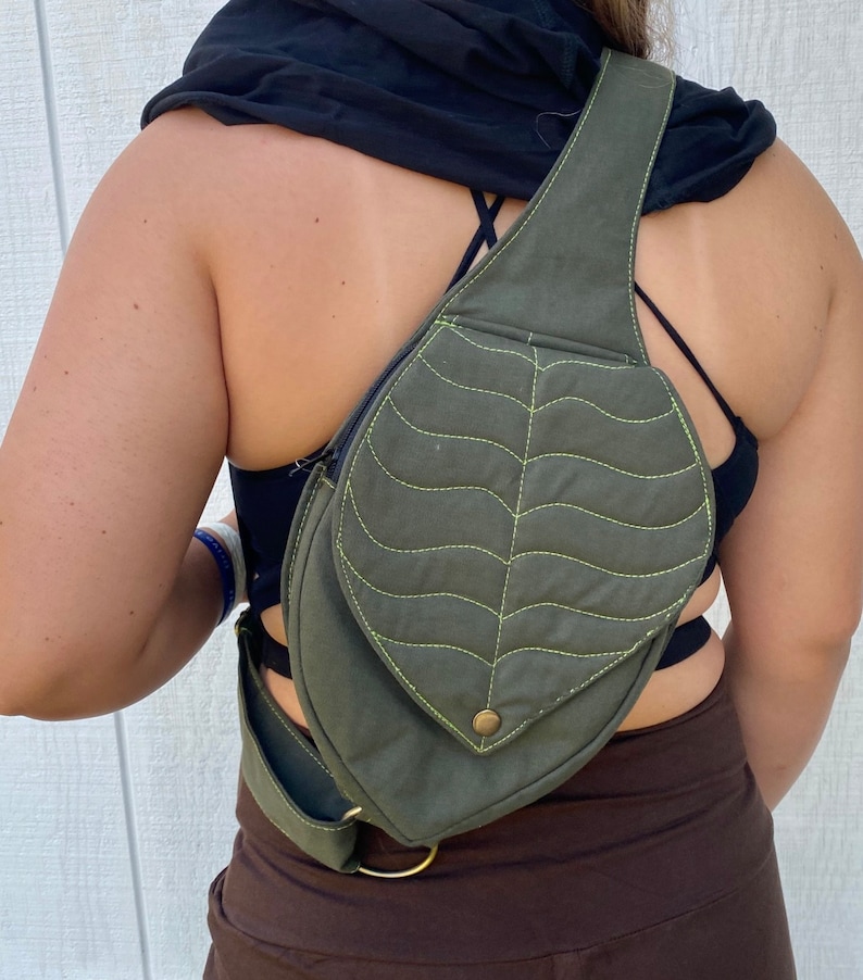 May include: A green canvas crossbody bag with a leaf-shaped flap and a yellow stitch detail. The bag has a strap that goes over the shoulder and a buckle on the side.