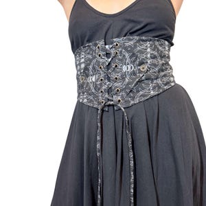 May include: A black corset belt with a white, abstract pattern. The belt features a lace-up front with metal grommets and decorative buttons. The corset is worn over a black dress.