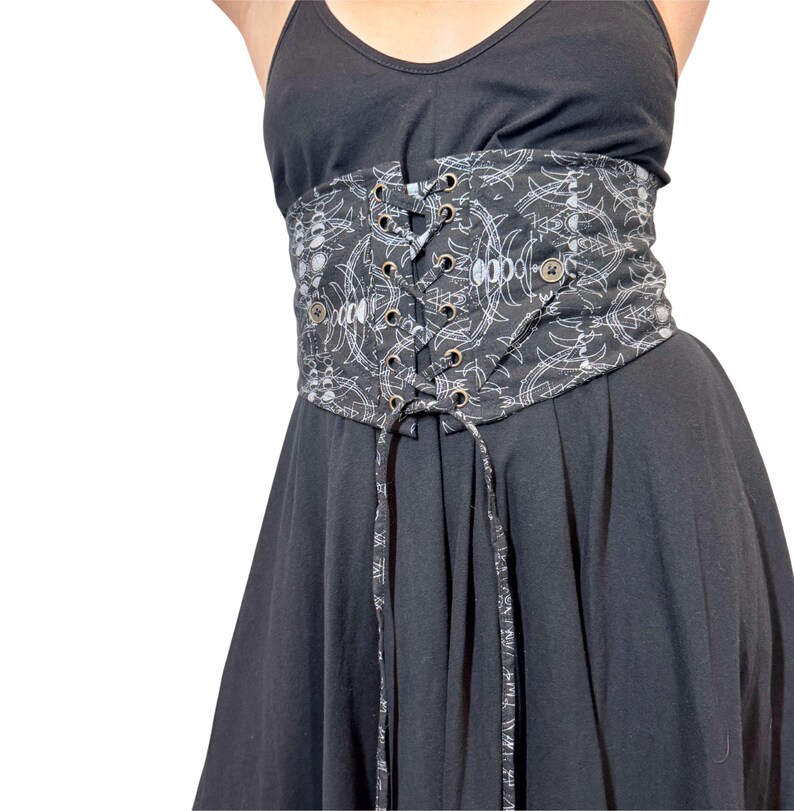 May include: A black corset belt with a white, abstract pattern. The belt features a lace-up front with metal grommets and decorative buttons. The corset is worn over a black dress.
