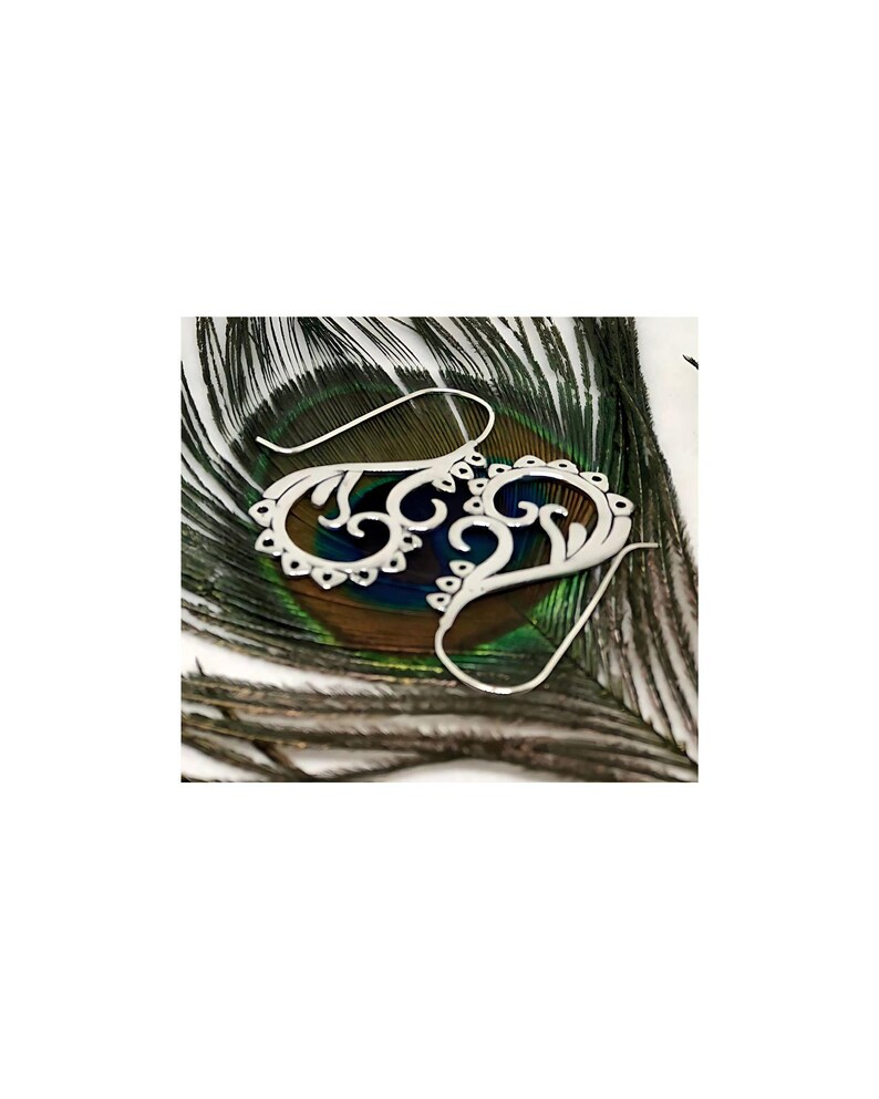 May include: A pair of silver filigree earrings with a delicate, intricate design. The earrings are shown on a peacock feather background.