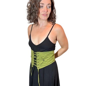 May include: A light green corset with a lace-up front and a pocket detail. The corset is worn over a black dress. The corset is made of a sturdy fabric and has a structured design.