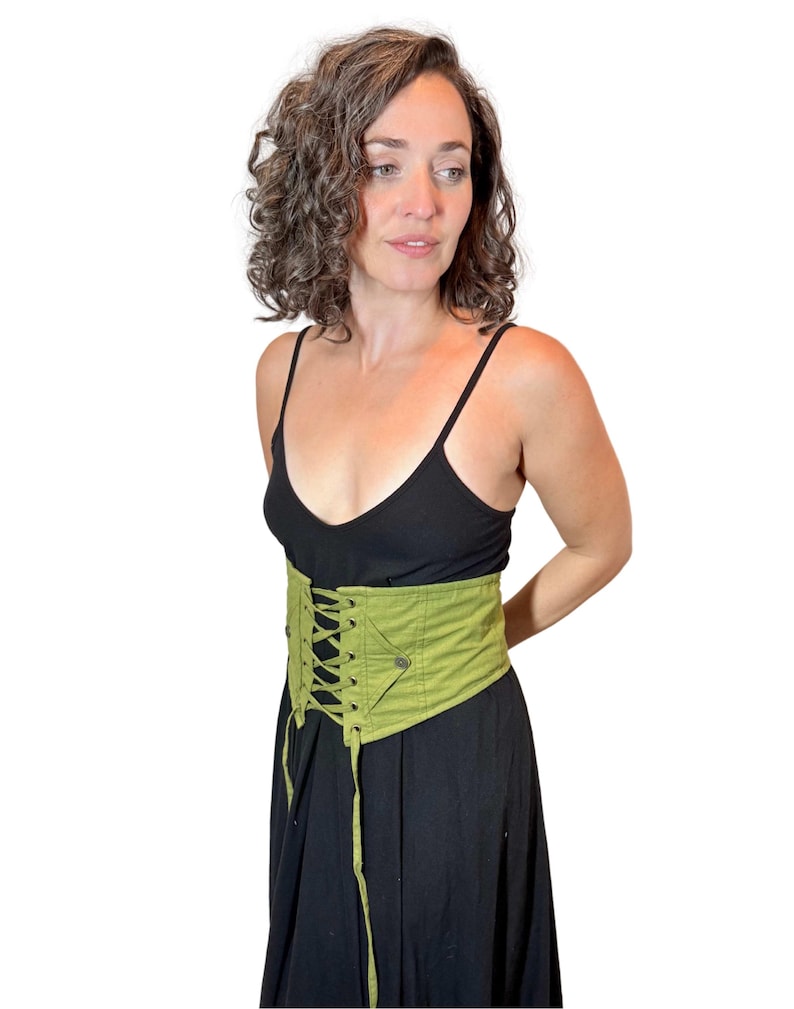May include: A light green corset with a lace-up front and a pocket detail. The corset is worn over a black dress. The corset is made of a sturdy fabric and has a structured design.
