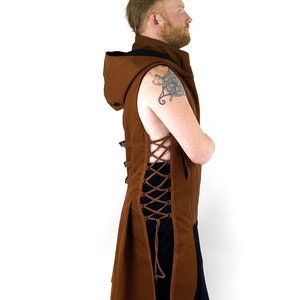 K&ouml;nnte beinhalten: Eine braune &auml;rmellose Weste mit Kapuze und langem, flie&szlig;endem R&uuml;cken. Die Weste hat ein Schn&uuml;rdetail auf der R&uuml;ckseite und ist aus einem strukturierten Stoff gefertigt.