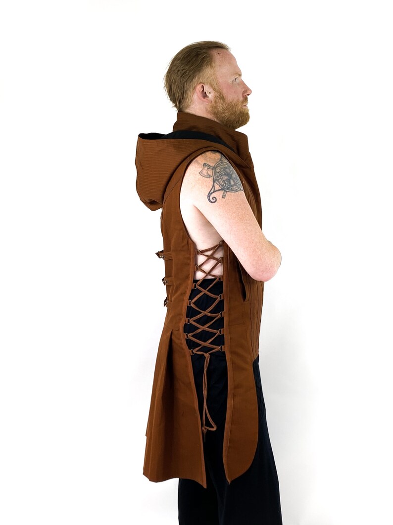 K&ouml;nnte beinhalten: Eine braune &auml;rmellose Weste mit Kapuze und langem, flie&szlig;endem R&uuml;cken. Die Weste hat ein Schn&uuml;rdetail auf der R&uuml;ckseite und ist aus einem strukturierten Stoff gefertigt.