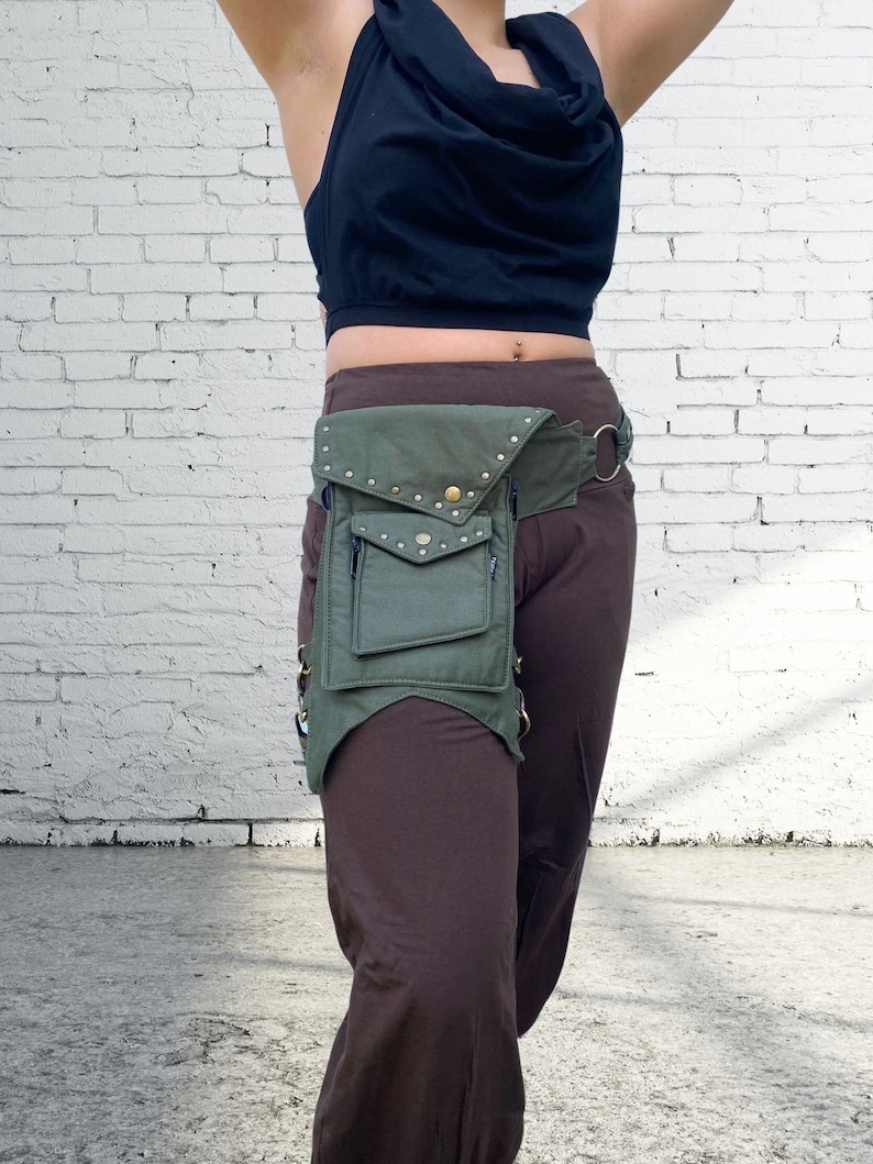 Puede incluir: Una ri&ntilde;onera de lona verde con varios bolsillos y detalles met&aacute;licos. La ri&ntilde;onera se lleva sobre un pantal&oacute;n marr&oacute;n.