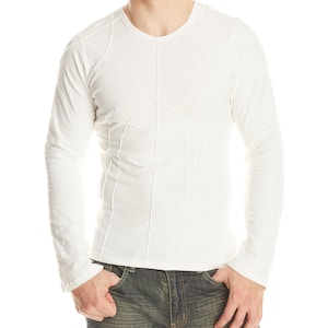 May include: A long-sleeved, off-white shirt with a V-neck and decorative stitching. The shirt has a fitted design and is paired with blue jeans. The model is standing with their hands in their pockets.