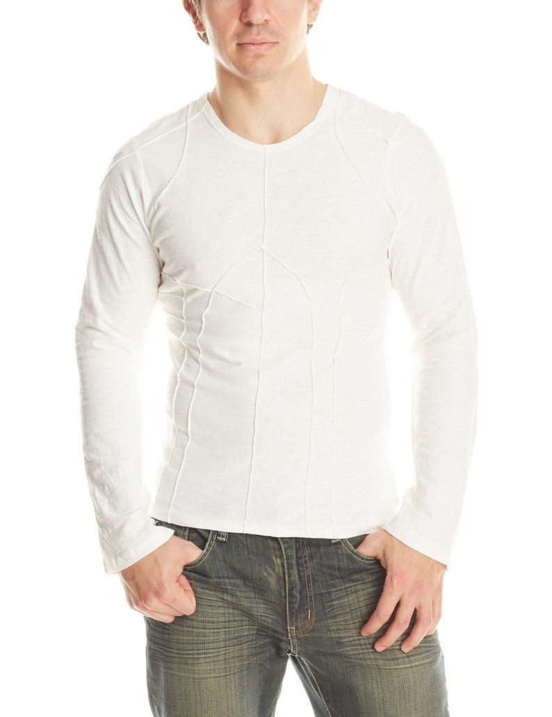 May include: A long-sleeved, off-white shirt with a V-neck and decorative stitching. The shirt has a fitted design and is paired with blue jeans. The model is standing with their hands in their pockets.
