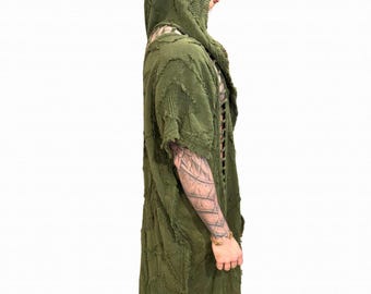 Puede incluir: Bata con capucha verde oliva con un diseño texturizado y desgastado. La bata presenta mangas cortas y una silueta larga y fluida. La tela tiene una apariencia única, similar a un patchwork. La prenda se usa sobre una camisa y pantalones oscuros.