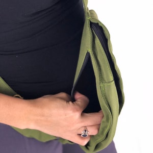 May include: A green canvas crossbody bag with a zippered pocket. The bag is worn by a person and the zipper is open, revealing the black lining.