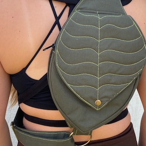 May include: A green canvas leaf-shaped crossbody bag with a yellow stitch detail and a brass buckle. The bag has a strap that can be adjusted to fit comfortably across the shoulder.