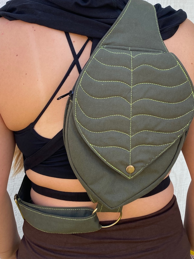 May include: A green canvas leaf-shaped crossbody bag with a yellow stitch detail and a brass buckle. The bag has a strap that can be adjusted to fit comfortably across the shoulder.