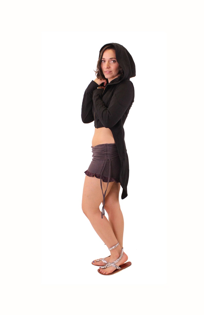 May include: A black hooded jacket with a long, flowing back. The jacket is cropped at the waist and has a drawstring hood. The model is wearing a gray skirt and sandals.