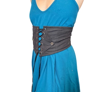 May include: A black corset belt with a lace-up front and a button closure. The belt is worn over a blue dress. The corset belt is made of a sturdy material and has a structured design.