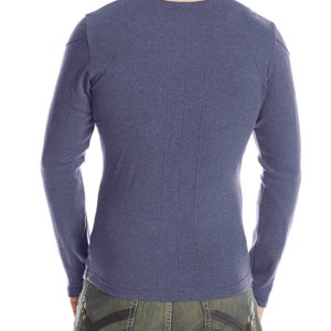 May include: A long-sleeved, blue shirt with a crew neck and vertical stitching down the back. The shirt is paired with blue jeans with a black design on the back pockets. The model is facing away from the camera.