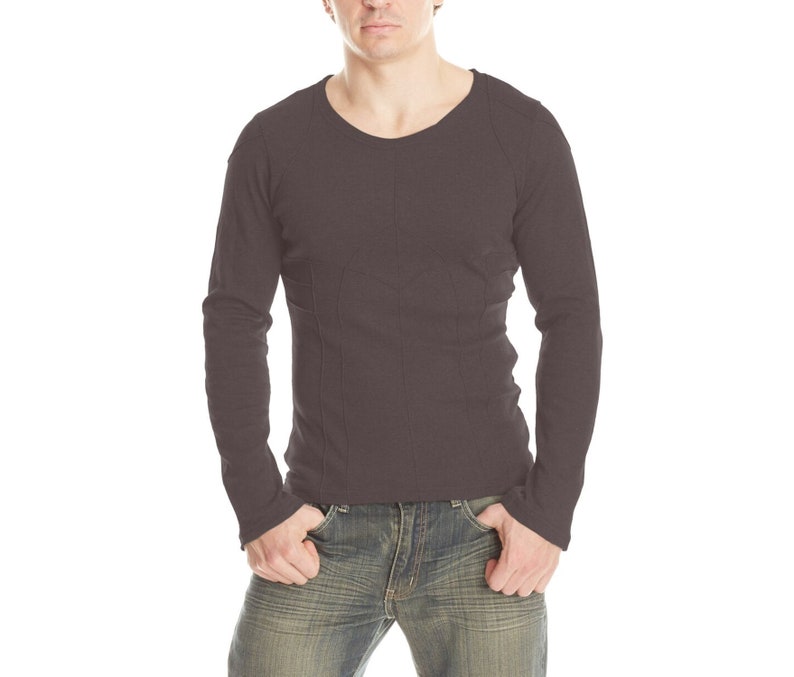 May include: A dark brown long-sleeve shirt with vertical stitching details. The shirt has a crew neck and is paired with blue jeans. The model is standing with their hands in their pockets.