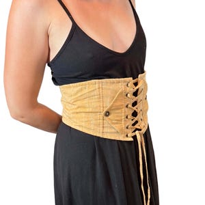 May include: A tan corset belt with a lace-up front and button closure. The belt is made of a textured fabric and is worn over a black dress. The corset belt cinches the waist and adds a unique style.