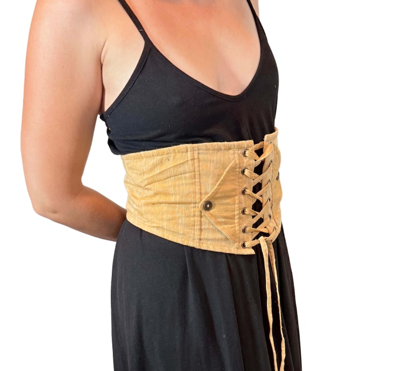 May include: A tan corset belt with a lace-up front and button closure. The belt is made of a textured fabric and is worn over a black dress. The corset belt cinches the waist and adds a unique style.