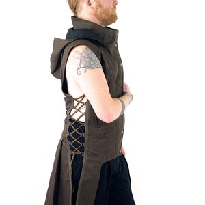 K&ouml;nnte beinhalten: Eine braune Kapuzenweste mit langem Schwanz und einem Schn&uuml;rdetail an der Seite. Die Weste ist aus einem strukturierten Stoff gefertigt und hat einen schwarzen Kragen.