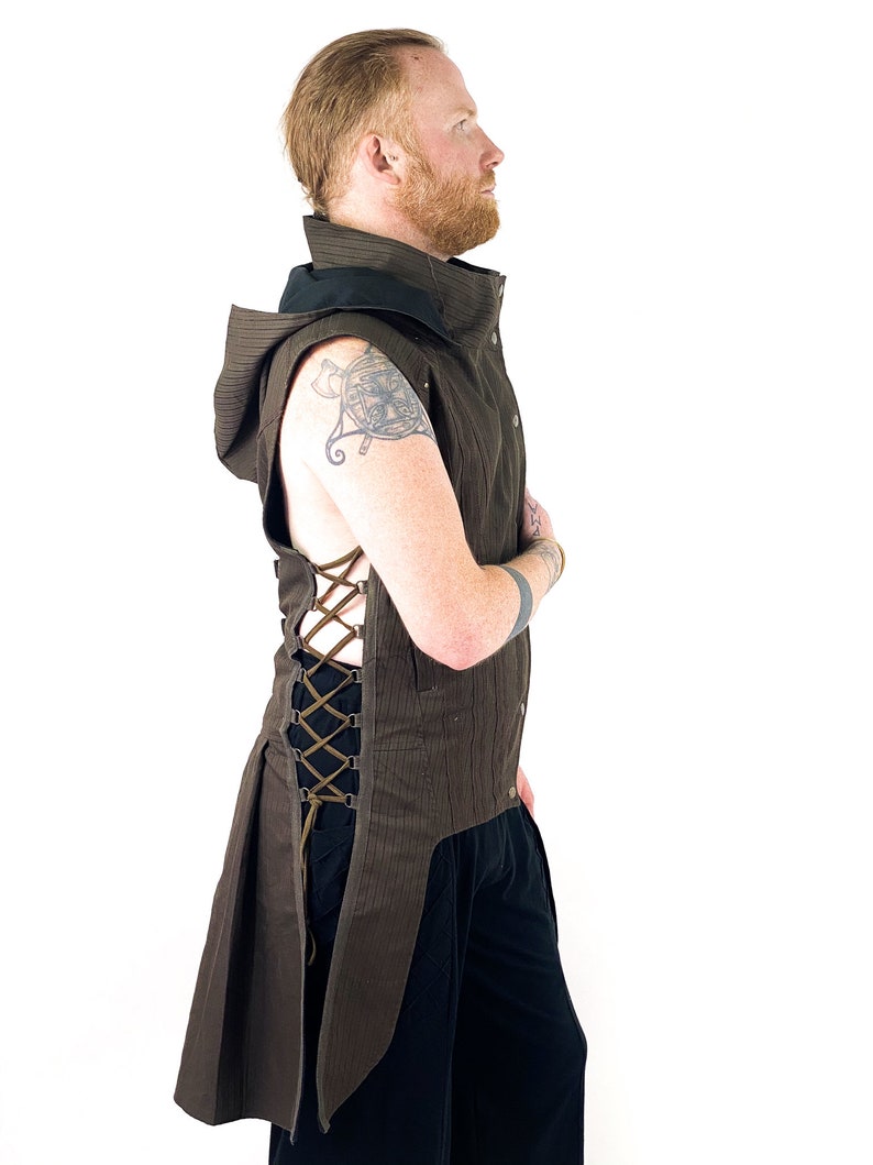 K&ouml;nnte beinhalten: Eine braune Kapuzenweste mit langem Schwanz und einem Schn&uuml;rdetail an der Seite. Die Weste ist aus einem strukturierten Stoff gefertigt und hat einen schwarzen Kragen.