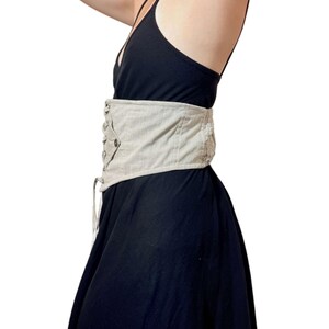 May include: A beige corset belt with a lace-up front and button details. The belt is worn over a black dress. The corset belt cinches the waist and adds a decorative element to the outfit.