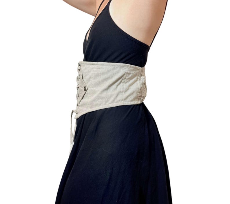May include: A beige corset belt with a lace-up front and button details. The belt is worn over a black dress. The corset belt cinches the waist and adds a decorative element to the outfit.