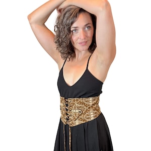 May include: A brown and beige patterned corset belt with a lace-up front is worn over a black dress. The corset belt is the focal point, featuring a detailed design. The overall style suggests a bohemian or vintage aesthetic.