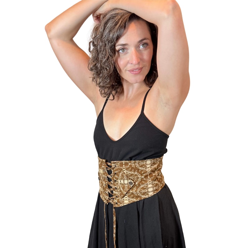 May include: A brown and beige patterned corset belt with a lace-up front is worn over a black dress. The corset belt is the focal point, featuring a detailed design. The overall style suggests a bohemian or vintage aesthetic.