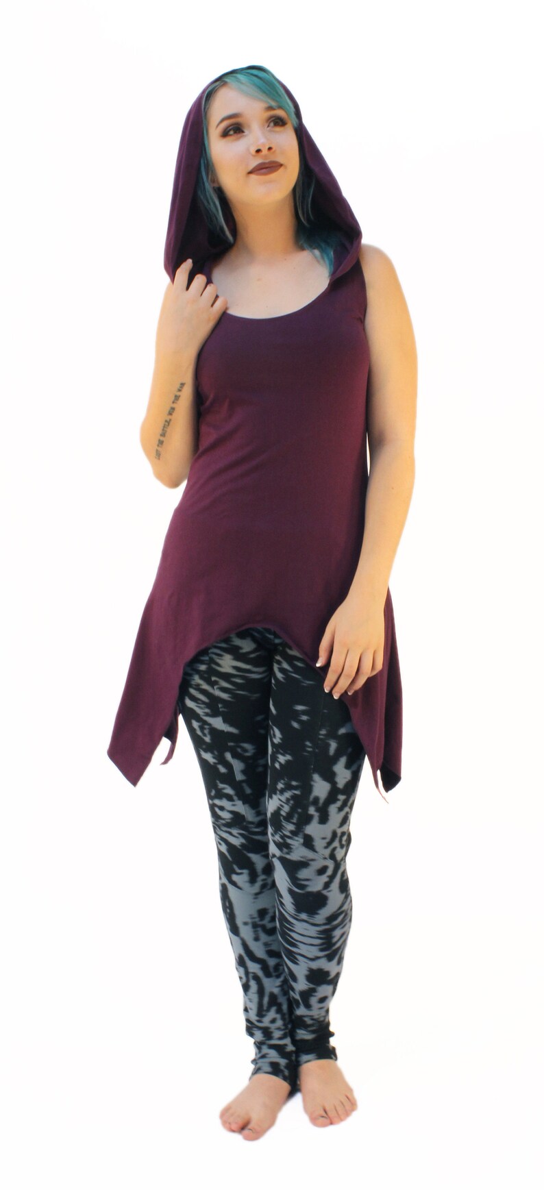 May include: A woman wearing a purple hooded tunic with a high-low hemline and black and gray patterned leggings. She is barefoot and has teal blue hair.