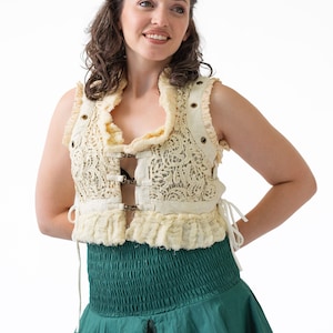 Op de afbeelding: Een vrouw draagt een cr&egrave;mekleurige kantvest met een groene rok. Het vest heeft een gerimpelde halslijn en is in de taille getailleerd met een gesp. De rok is een eenvoudige, vloeiende stijl.