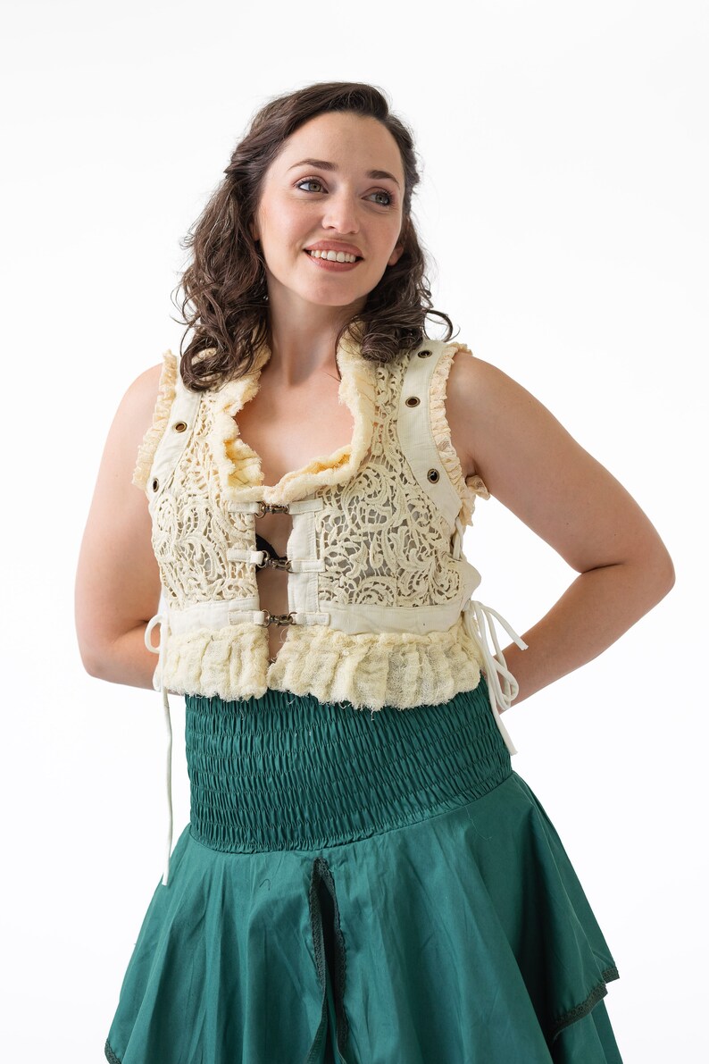 Op de afbeelding: Een vrouw draagt een cr&egrave;mekleurige kantvest met een groene rok. Het vest heeft een gerimpelde halslijn en is in de taille getailleerd met een gesp. De rok is een eenvoudige, vloeiende stijl.