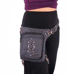 May include: A gray canvas thigh bag with a zippered pocket, a metal ring, and silver studs. The bag is worn on a person's right thigh and is secured with a belt.