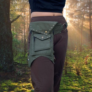 Puede incluir: Una ri&ntilde;onera de lona verde con tachuelas met&aacute;licas y un bolsillo con cremallera. La ri&ntilde;onera se lleva en un cintur&oacute;n marr&oacute;n con una hebilla met&aacute;lica. La persona que lleva la ri&ntilde;onera est&aacute; de pie en un bosque.