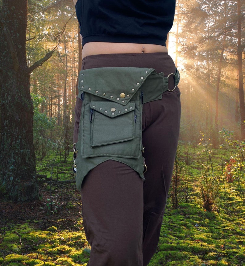 Puede incluir: Una ri&ntilde;onera de lona verde con tachuelas met&aacute;licas y un bolsillo con cremallera. La ri&ntilde;onera se lleva en un cintur&oacute;n marr&oacute;n con una hebilla met&aacute;lica. La persona que lleva la ri&ntilde;onera est&aacute; de pie en un bosque.