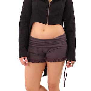 May include: A black hooded cropped jacket with a long back tail and a zipper closure. The jacket is worn over a pair of dark brown shorts with a ruffled hem.
