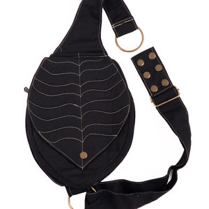 May include: A black canvas crossbody bag with a unique leaf-shaped design and a decorative stitched pattern. The bag has a long adjustable strap with brass hardware and a small zippered pocket.