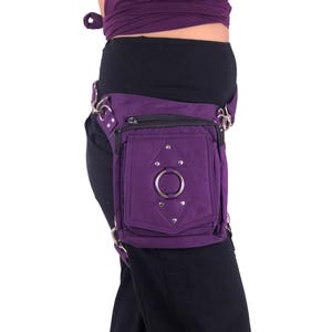 May include: Purple canvas thigh bag with a silver ring and silver studs. The bag has a zipper closure and a small pocket on the front.