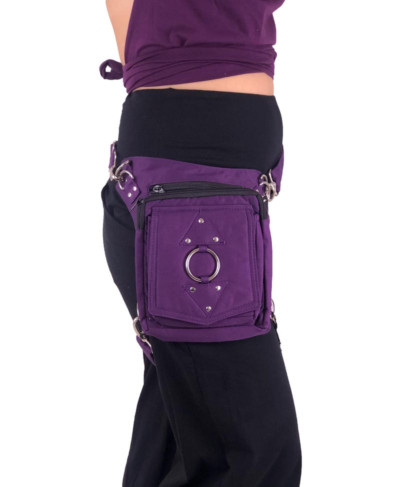 May include: Purple canvas thigh bag with a silver ring and silver studs. The bag has a zipper closure and a small pocket on the front.