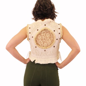 Op de afbeelding: Een wit gehaakt vest met een achterpaneel van kant en een bruine leren bies. Het vest heeft een getailleerde lengte en een gerimpelde zoom.
