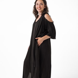 May include: A black, open-shoulder maxi dress with a V-neck and short sleeves. The dress has a relaxed fit and a raw hemline. The model is smiling and standing in front of a white background. The dress has pockets.