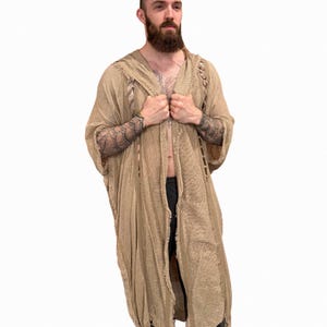 May include: A beige hooded robe with long sleeves and a distressed texture. The robe has a loose fit and a frayed hemline. The sleeves are wide, and the robe is open at the front. The garment is made of a lightweight fabric.
