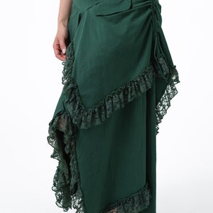 Puede incluir: Una falda larga de color verde oscuro con ribete de encaje. La falda tiene un detalle fruncido en la parte posterior y un bolsillo en el lado derecho.