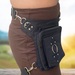 May include: A black canvas fanny pack with a zippered pocket and a large metal ring. The fanny pack is worn on the hip and has a strap that goes around the leg.