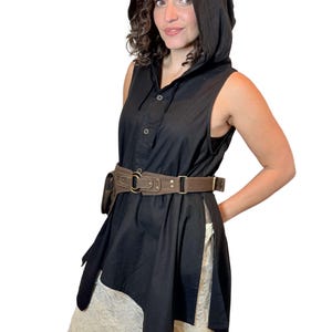 May include: Black sleeveless hooded tunic with a button-down front and asymmetrical hem. A brown leather belt with a pouch and metal buckle cinches the waist. The outfit is paired with a cream-colored skirt.
