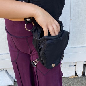 Może przedstawiać: Czarna sk&oacute;rzana torebka na udo z zamkiem błyskawicznym i paskiem, kt&oacute;ry owija się wok&oacute;ł talii. Torebka jest noszona na prawym udzie osoby w fioletowej sp&oacute;dnicy.