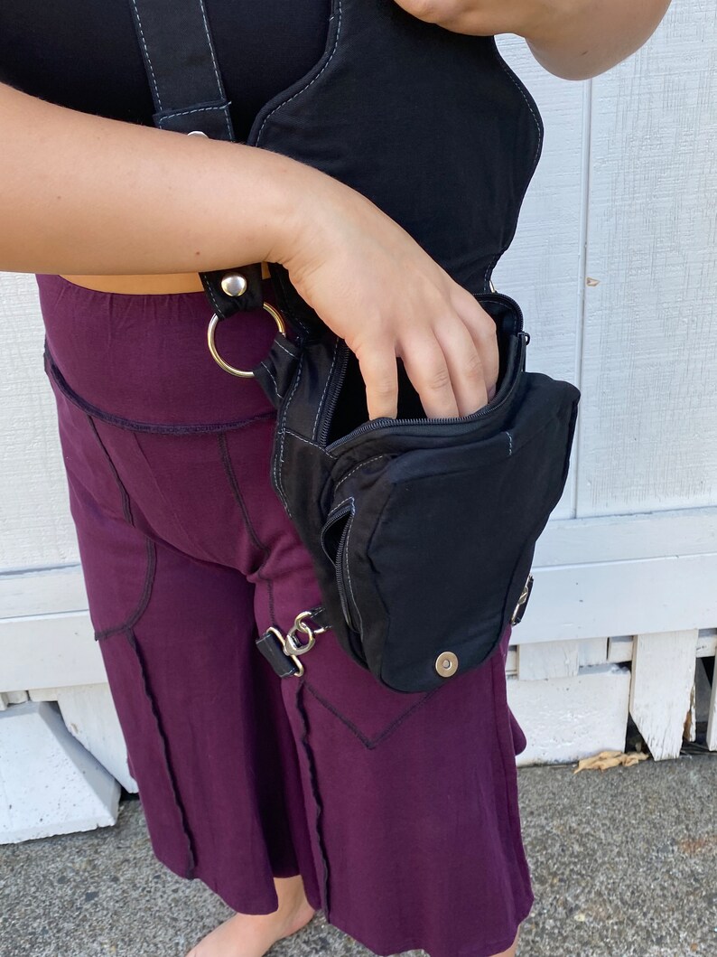 Może przedstawiać: Czarna sk&oacute;rzana torebka na udo z zamkiem błyskawicznym i paskiem, kt&oacute;ry owija się wok&oacute;ł talii. Torebka jest noszona na prawym udzie osoby w fioletowej sp&oacute;dnicy.